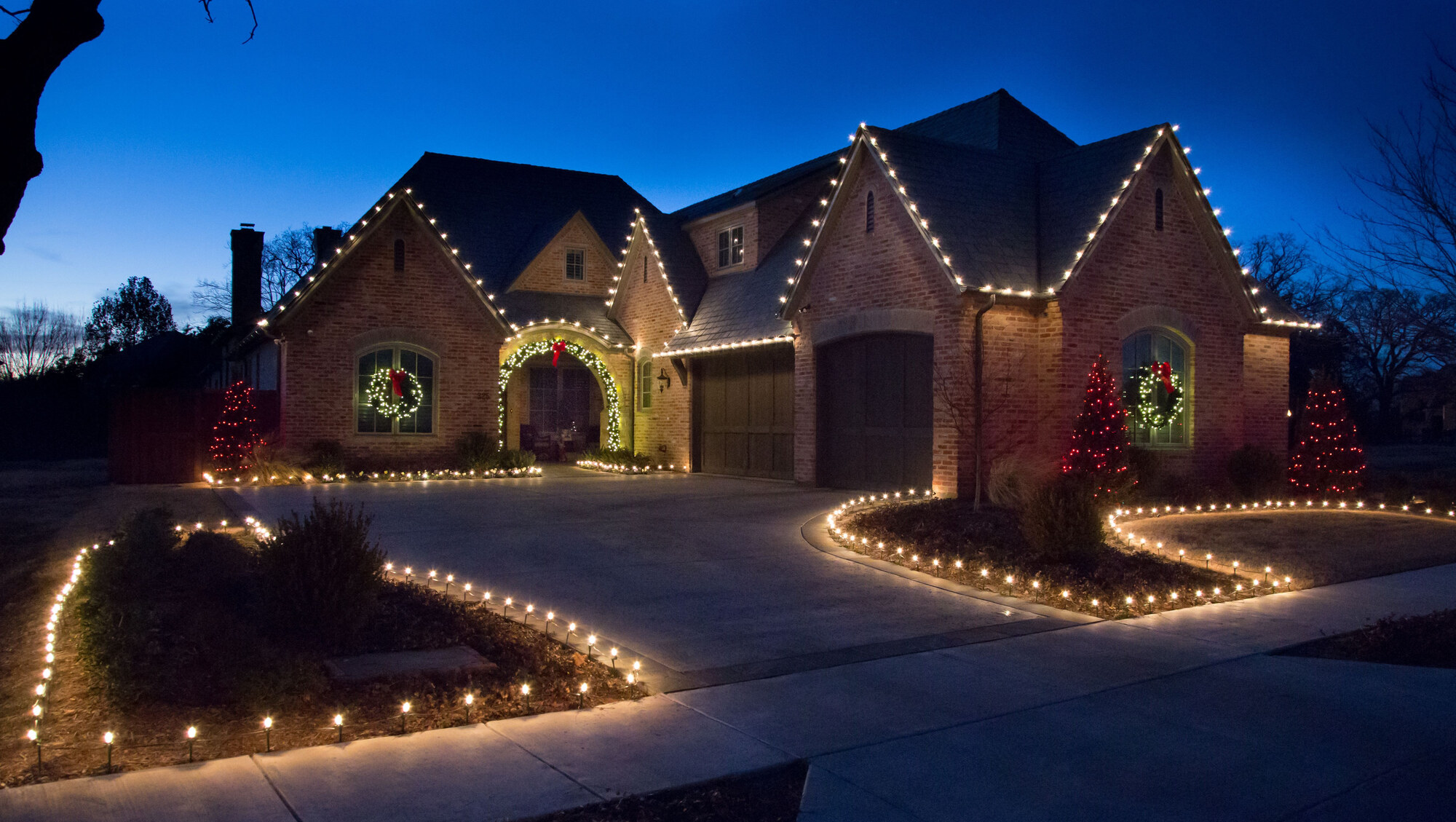 Brick home with warm white roofline lights, wreaths, and driveway stake lights