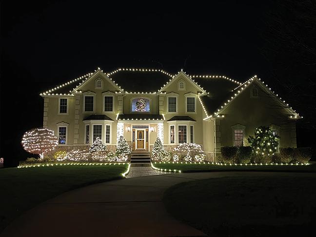 Two-story home with warm white Christmas lights, wreath, and wrapped shrubs