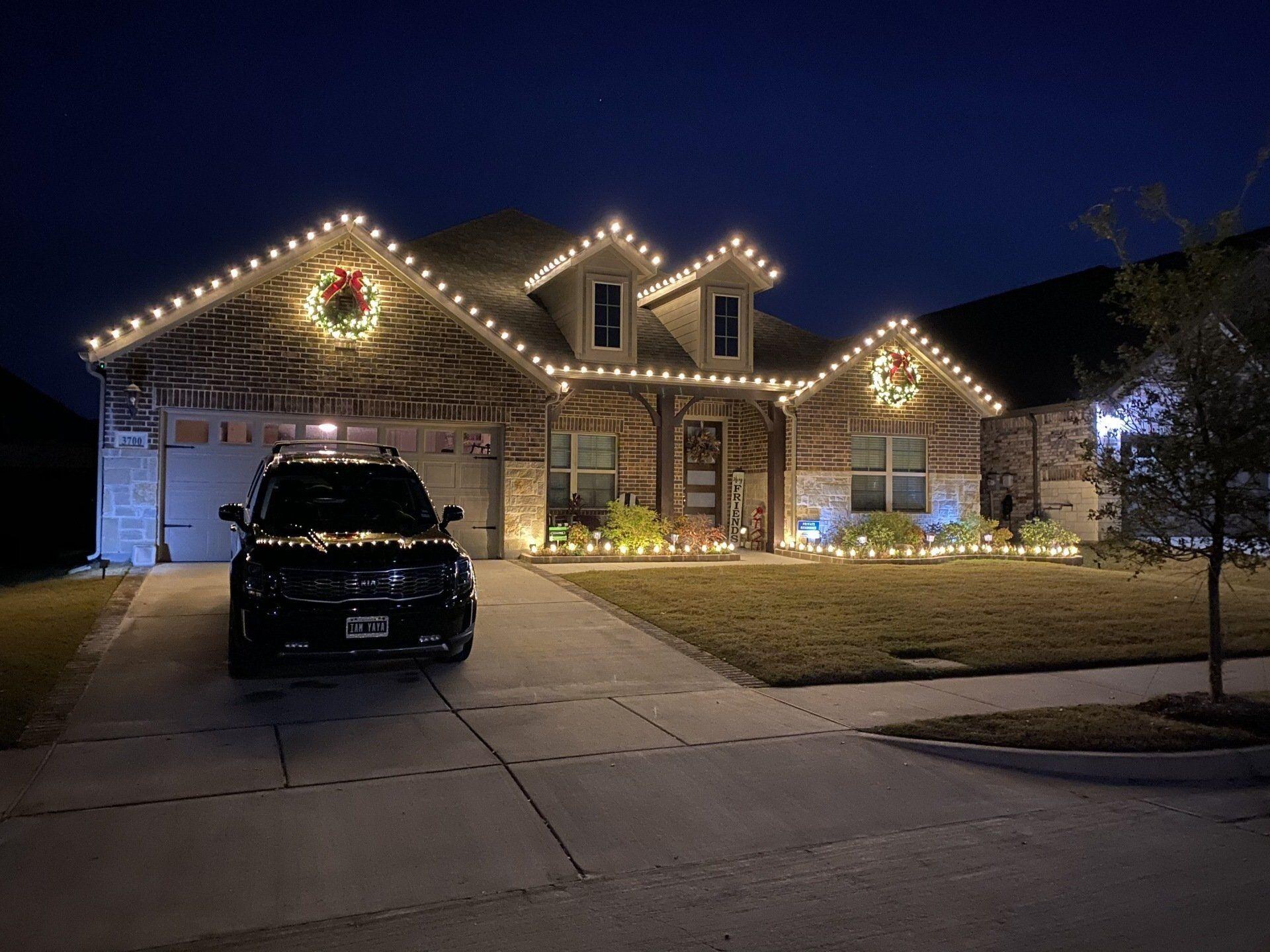 Brick home with warm white roofline lights, lit wreaths, and garden bed lights